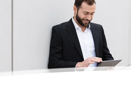 Das Bild zeigt einen Mann in einem Anzug, der an einer Wand lehnt und auf ein Tablet schaut. Er trägt ein weißes Hemd und ein dunkles Jacket und lächelt, während er das Gerät bedient. Die Wand im Hintergrund ist schlicht und weiß, was dem Bild einen minimalistischen Look verleiht.