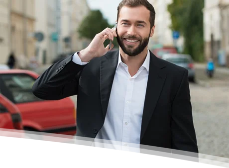 Das Bild zeigt einen Mann mit einem dunklen Bart, der ein weißes Hemd und einen schwarzen Blazer trägt. Er steht auf einer belebten Stadtstraße und lächelt, während er in ein schwarzes Handy spricht. Im Hintergrund sind verschwommene Autos und Gebäude zu sehen, was darauf hindeutet, dass es sich um eine lebhafte städtische Umgebung handelt. Das Licht scheint freundlich und die Atmosphäre wirkt positiv und energiegeladen. Der Mann strahlt Freude und Selbstbewusstsein aus, was das Bild besonders ansprechend macht.