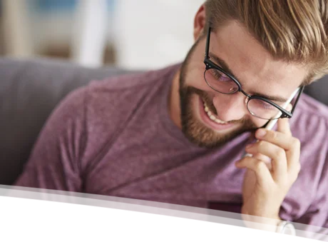 Das Bild zeigt einen jungen Mann mit Brille und Bart, der lächelnd am Telefon sitzt. Er hat blonde Haare und trägt ein purpurfarbenes T-Shirt. Der Hintergrund ist unscharf, aber es scheint sich um ein Wohnzimmer zu handeln.  Der Fokus liegt auf dem Mann und seinem freundlichen Gesichtsausdruck.