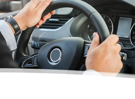 Das Bild zeigt eine Nahaufnahme von Händen, die fest das Lenkrad eines Autos halten. Der Lenkrad ist aus schwarzem Leder und wirkt modern und hochwertig. Die Hände des Fahrers sind gut sichtbar, wobei eine Hand den oberen Teil des Lenkrads umschließt, während die andere Hand etwas weiter unten greift.  Im Hintergrund sieht man einen Teil des Armaturenbretts des Fahrzeugs, das in einem eleganten Silberton gehalten ist. Es gibt digitale Anzeigen und Bedienelemente, die auf eine moderne Ausstattung des Autos hinweisen. Der Fahrer trägt ein weißes Hemd und hat ein Armband oder eine Uhr am Handgelenk, was einen gepflegten Eindruck vermittelt.  Die Atmosphäre des Bildes vermittelt ein Gefühl von Konzentration und Kontrolle, während der Fahrer das Auto lenkt. Es könnte eine Momentaufnahme einer Fahrt durch die Stadt oder auf einer Landstraße sein, die den Spaß und die Freiheit des Fahrens einfängt.