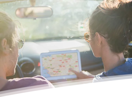 Das Bild zeigt zwei Personen, die in einem Cabrio sitzen und gemeinsam auf ein Tablet schauen, auf dem eine digitale Karte angezeigt wird. Die Person im Vordergrund, eine Frau mit hochgestecktem Haar, zeigt auf die Karte. Es scheint sich um eine gemeinsame Reiseplanung oder Orientierung während einer Fahrt zu handeln. Das Bild vermittelt ein Gefühl von Abenteuerlust, Freiheit und gemeinsamer Entdeckung. Die sonnige Atmosphäre verstärkt diesen Eindruck.