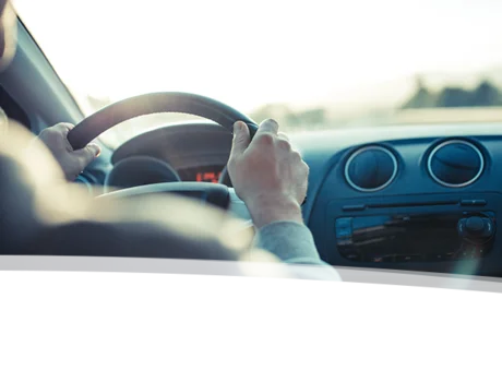 Das Bild zeigt die Hände einer Person am Steuer eines Autos. Der Fokus liegt auf den Händen und dem Lenkrad; der Rest des Autos und die Umgebung sind unscharf dargestellt. Das Licht deutet auf eine sonnige, möglicherweise frühe Morgen- oder späte Nachmittagsstimmung hin.  Es wirkt insgesamt ruhig und vermittelt ein Gefühl von Bewegung und Reise.