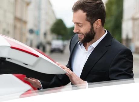 Das Bild zeigt einen Mann in einem Anzug, der an einem Auto steht und den Kofferraum öffnet. Er trägt einen gepflegten Bart und lächelt, während er sich auf den Kofferraum konzentriert. Im Hintergrund sind einige Gebäude und Bäume zu sehen, was auf eine städtische Umgebung hinweist.